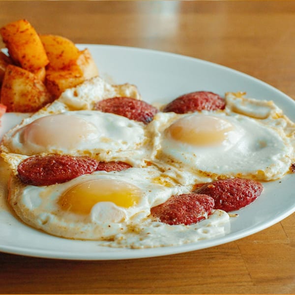 A Turkish breakfast plate with three fried eggs, sucuk sausage, and roasted potatoes.