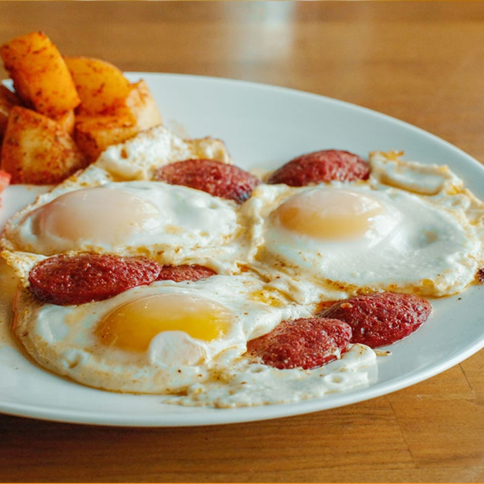 A Turkish breakfast plate with three fried eggs, sucuk sausage, and roasted potatoes.