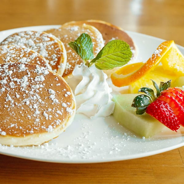 Fluffy pancakes dusted with powdered sugar, served with whipped cream, mint, strawberries, melon, and orange slices.