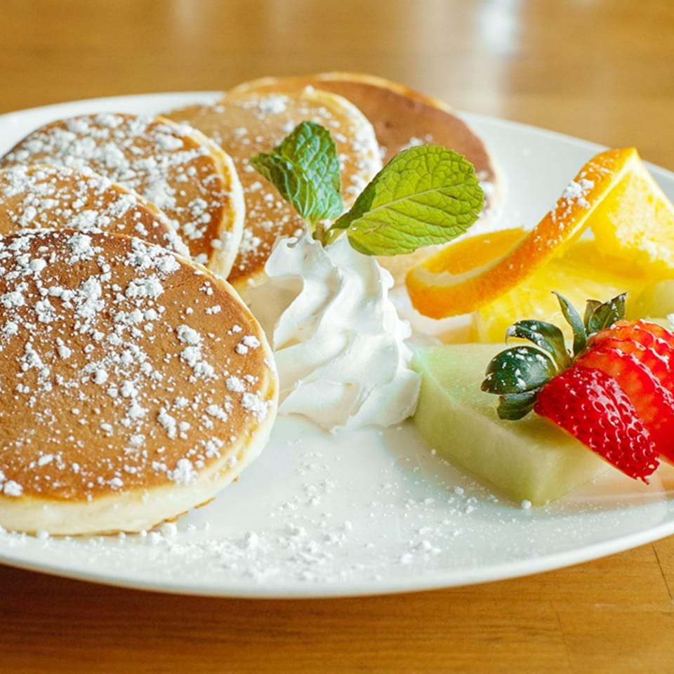 Fluffy pancakes dusted with powdered sugar, served with whipped cream, mint, strawberries, melon, and orange slices.