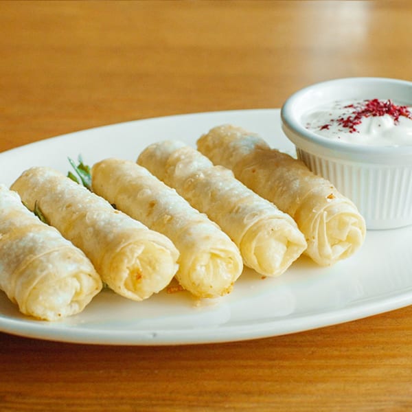 Five golden Turkish cigar borek pastries on a white plate with a side of yogurt dip.