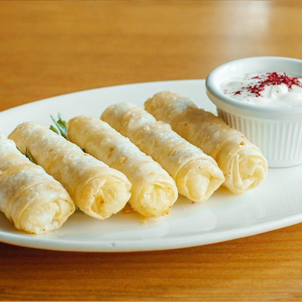 Five crispy Turkish cigar borek pastries with a side of yogurt dip, garnished with sumac and herbs.