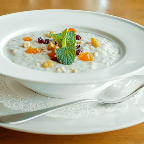 A white bowl of oatmeal with dried fruit and mint, next to a spoon on a doily.